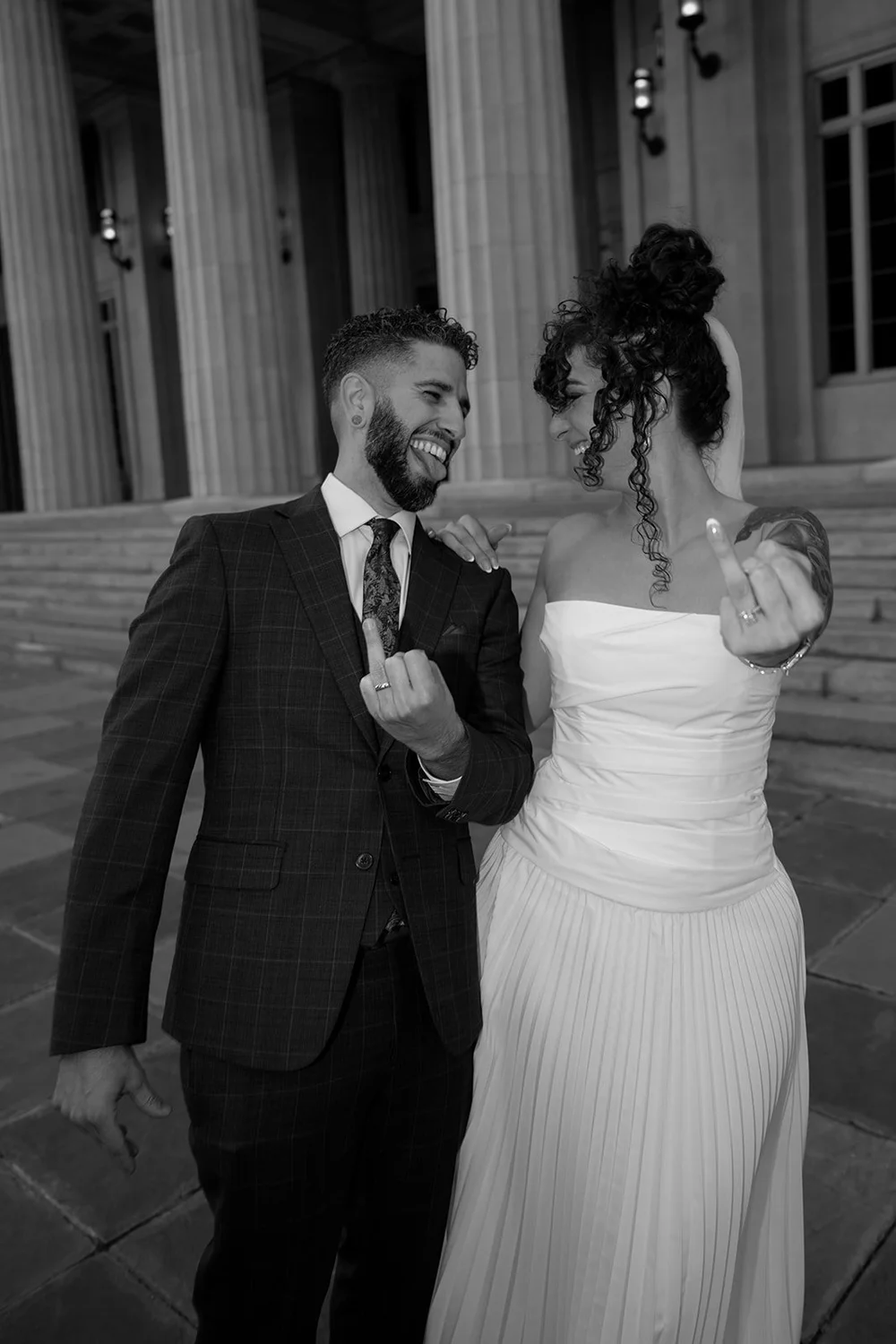Black and white photo of the groom laughing and pointing at the camera with the bride beside him in front of courthouse columns