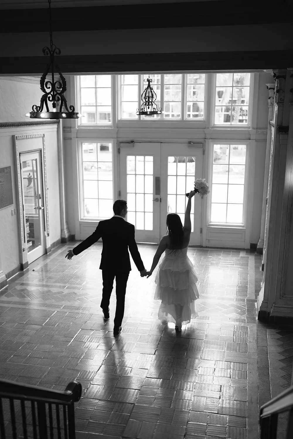 Black and white view from the stairs of the couple holding hands and walking toward tall glass doors