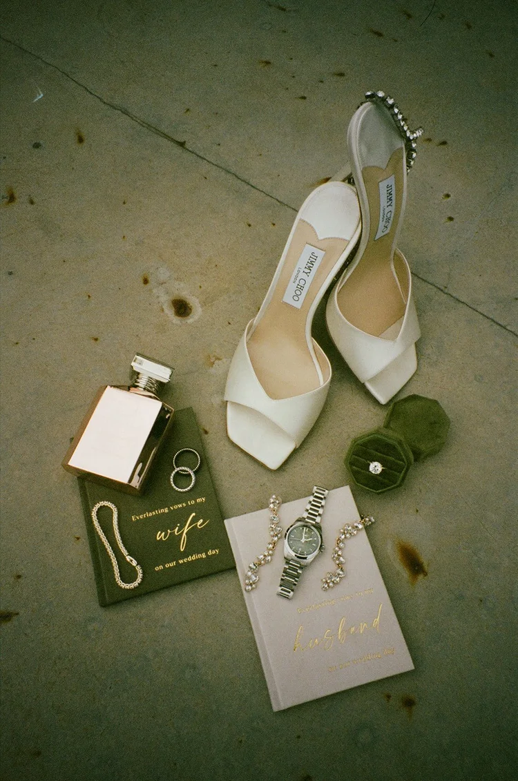 Film photo of white wedding heels, a perfume bottle, a watch and a vow book on a concrete floor