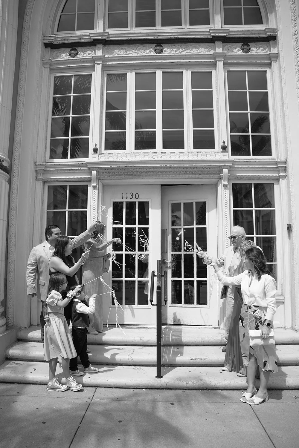 Black and white group photo of the wedding party on the steps in front of tall glass doors