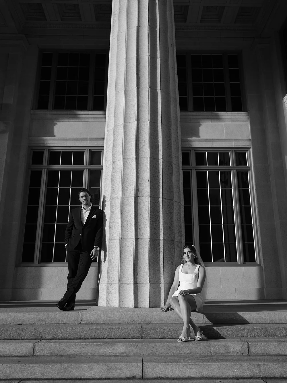 Black and white frame with the groom leaning on a column and the bride sitting on the steps below the courthouse windows