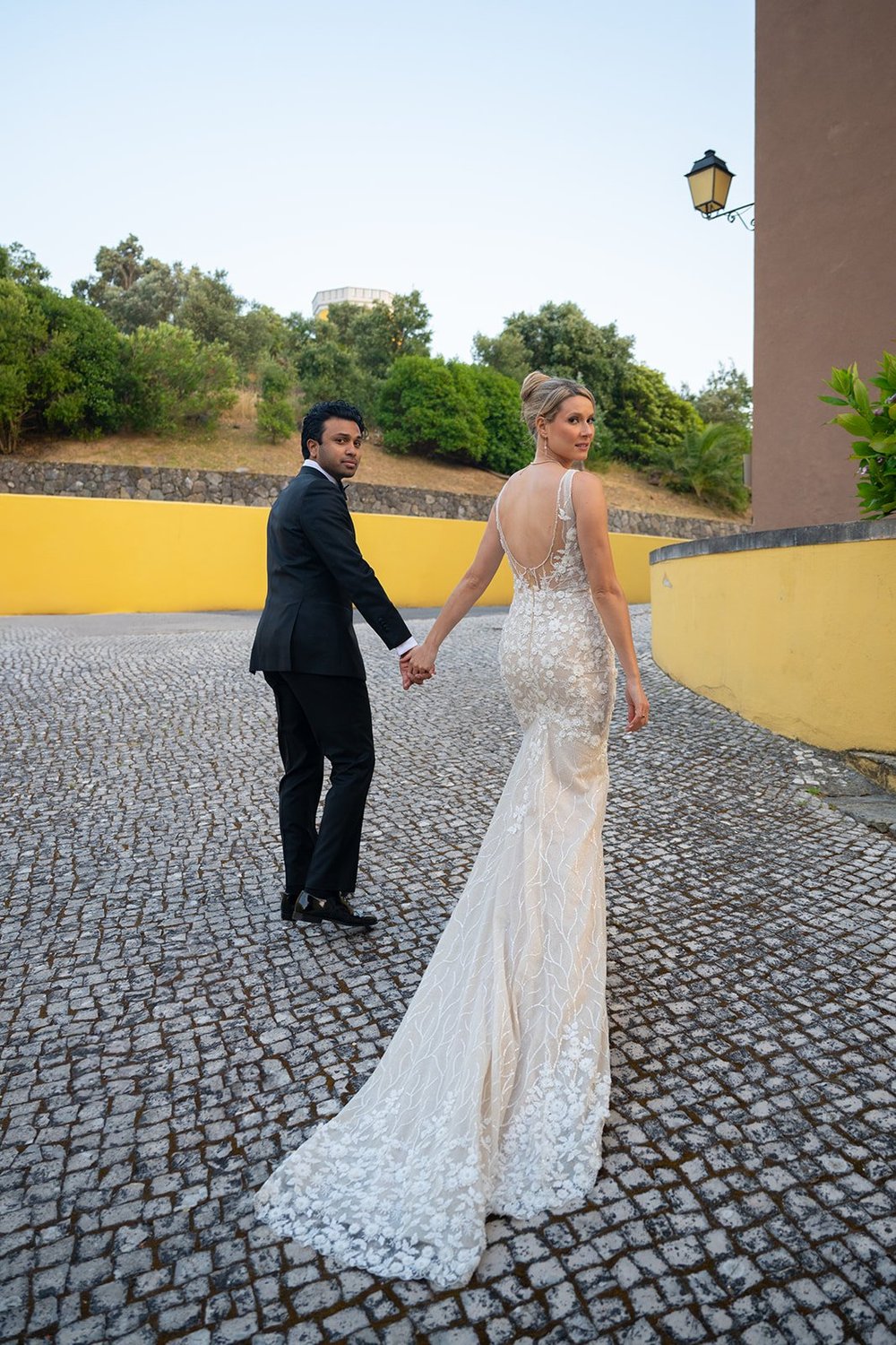 Ellen and Jacob walking hand in hand on a cobblestone path past curved yellow walls, Ellen looking back over her shoulder in her open-back gown