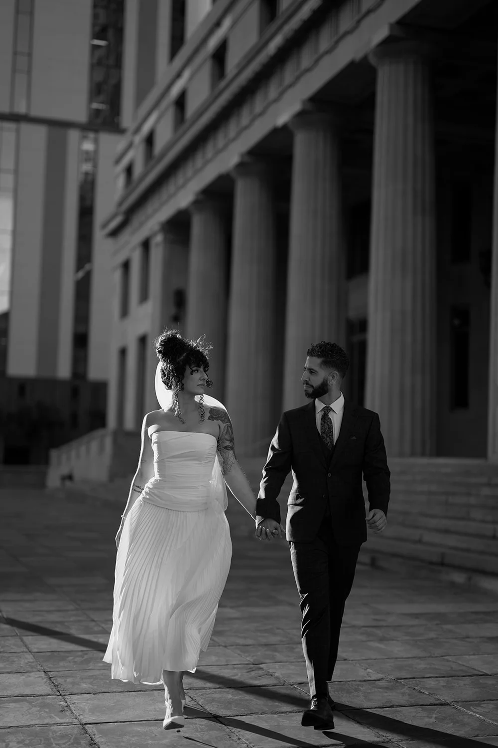 Black and white photo of the bride and groom walking hand in hand across a stone plaza in front of courthouse columns