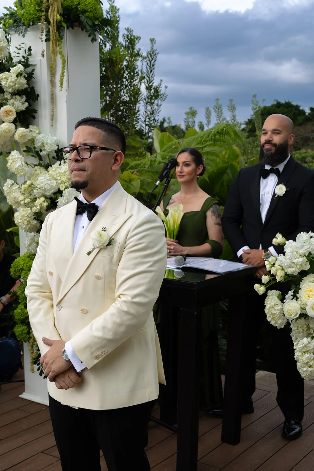 Douglas in a cream double-breasted jacket and bow tie waiting at the altar with his hands clasped, the officiant and a bridesmaid in green behind him
