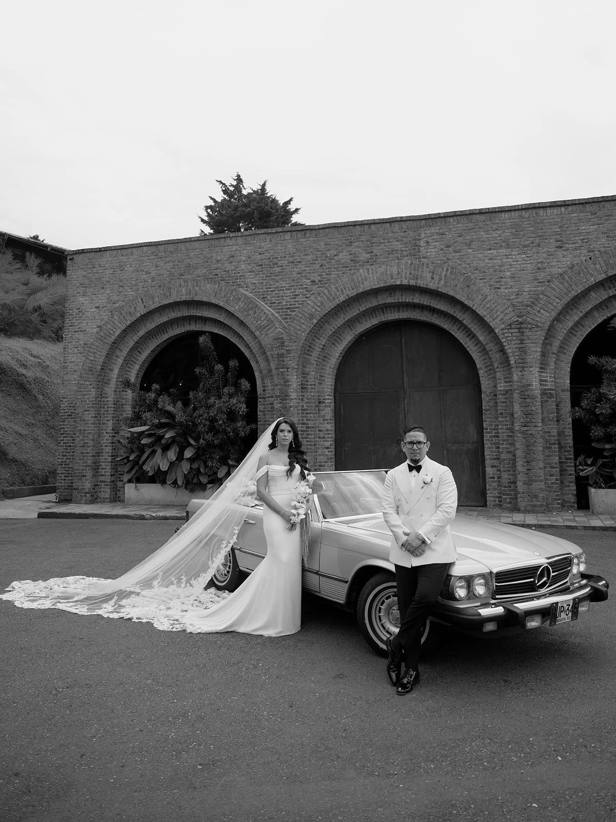 Black and white photo of Krystine and Douglas beside a vintage Mercedes convertible in front of brick arches at Voilà Eventos in Medellín