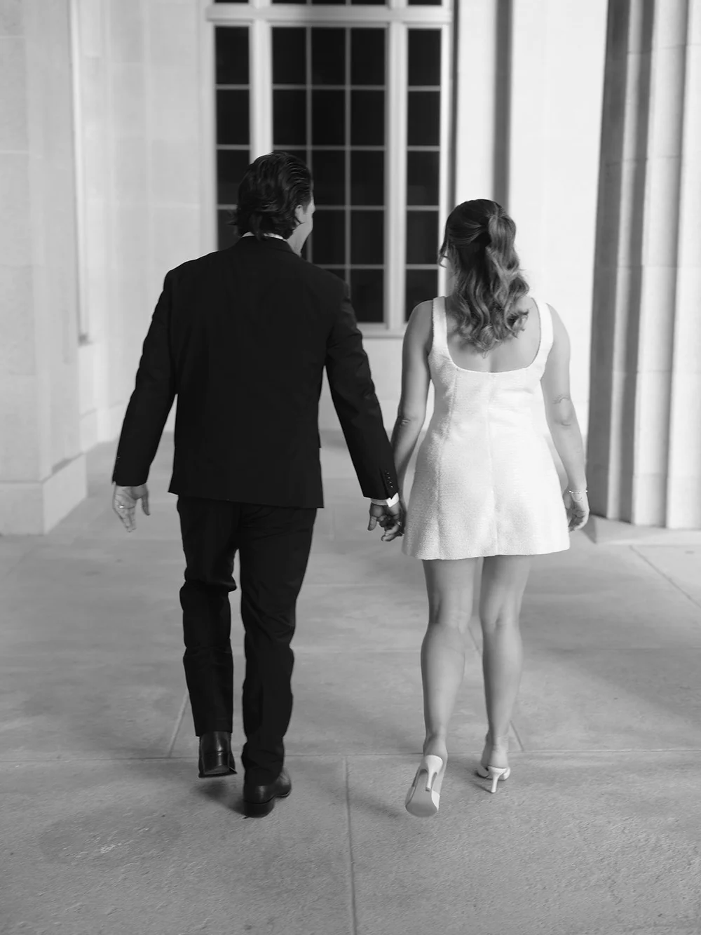 Black and white image from behind of the couple walking hand in hand along the courthouse walkway