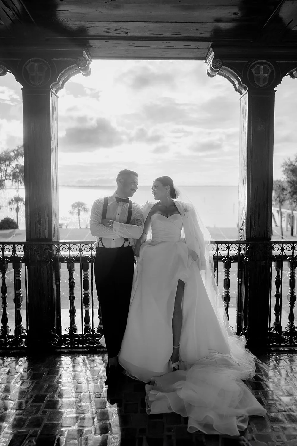 Black and white photo of Steph and Ricky talking on a covered terrace with carved wooden columns and the water behind them