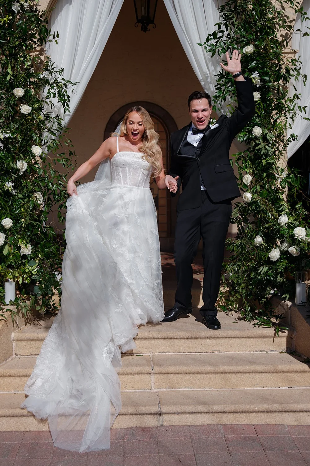 Jane smiling and Berkeley cheering with his arm raised on the steps in front of the ceremony arch