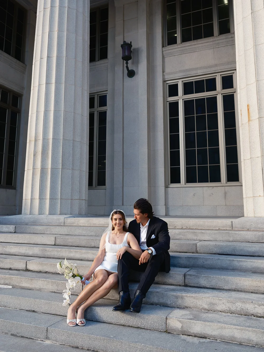 Carina and Billy sitting close on the wide stone courthouse steps with the columns above them and her white bouquet beside her