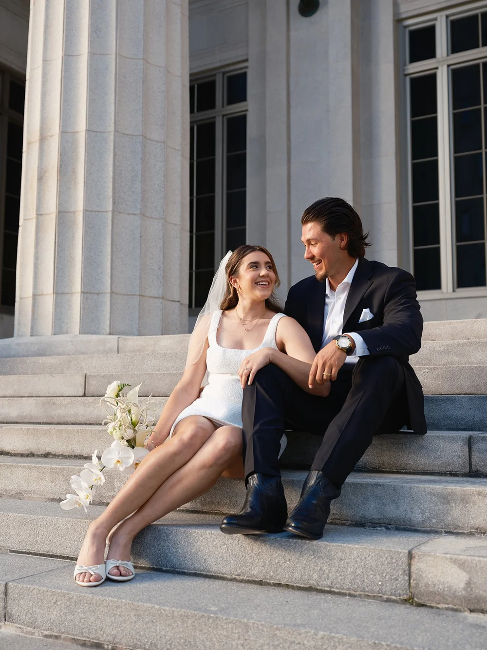 Bride in a short white dress and groom in a black suit sitting side by side on the courthouse steps, smiling