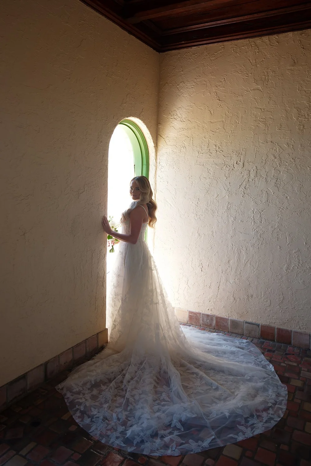Jane standing in a sunlit arched window with her long lace train spread on the floor