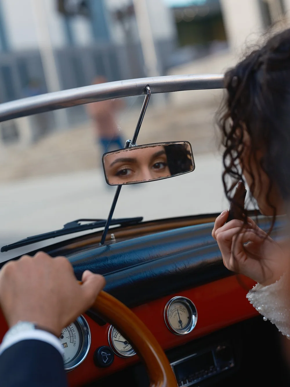 Detail of the groom's hand on the steering wheel and the bride's eye reflected in the rearview mirror