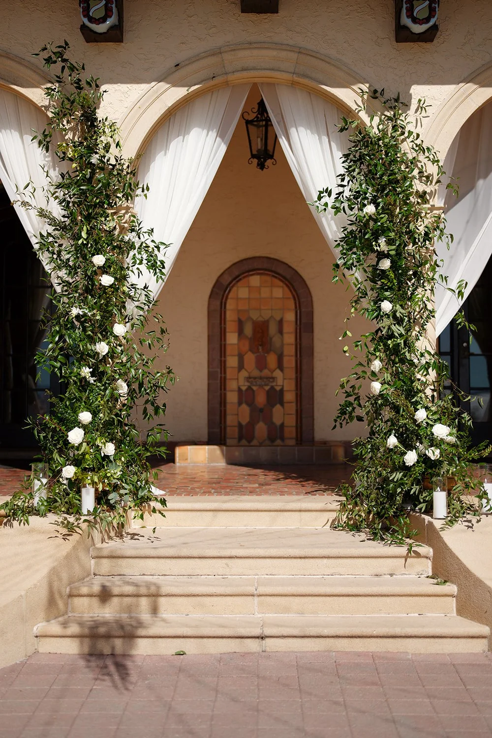 The ceremony arch at the Powel Crosley Estate dressed with greenery, white roses and white drapes in front of a carved wooden door