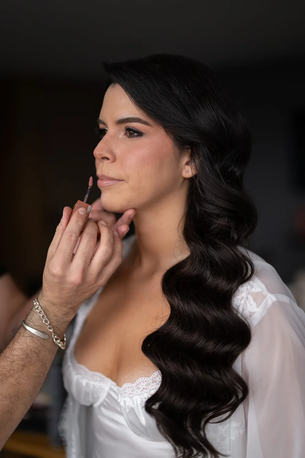 Krystine in a white robe touching her face while getting ready, her dark hair in long waves