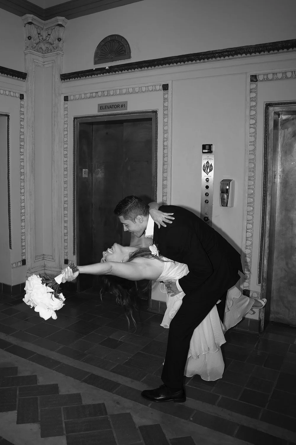 Black and white photo of David leaning over Chris in the hallway with her bouquet held out to the side