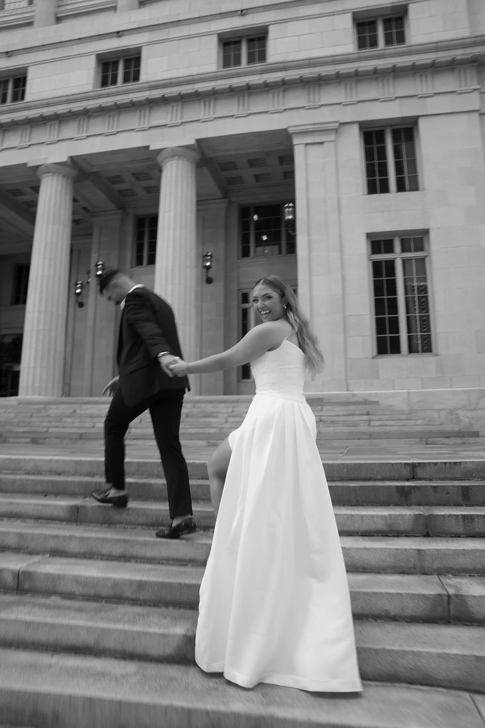 Rachel in a long white gown walking across the courthouse steps with Austin behind her, in black and white
