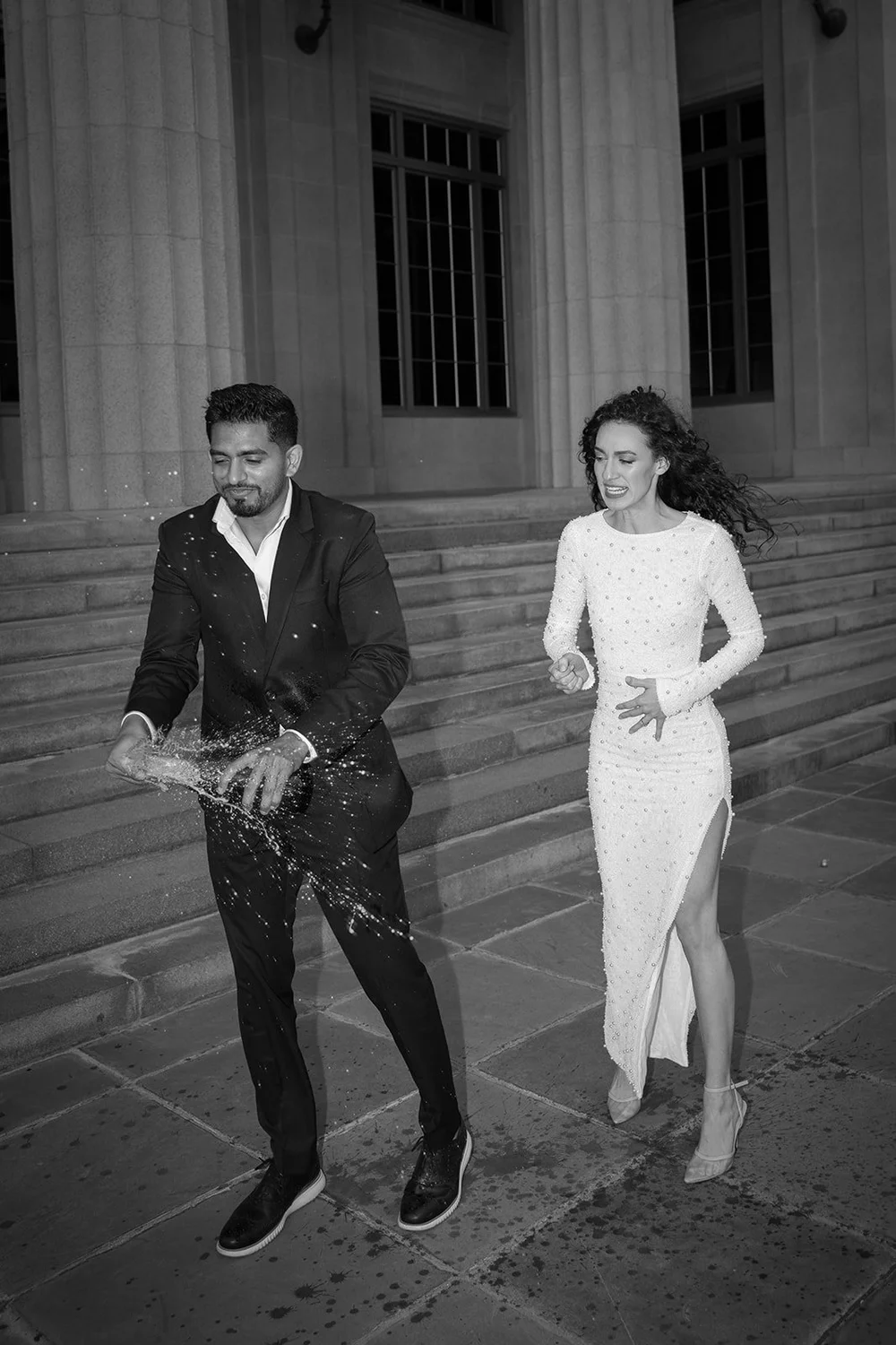 Black and white image of the groom spraying a bottle as the bride runs beside him across the courthouse steps