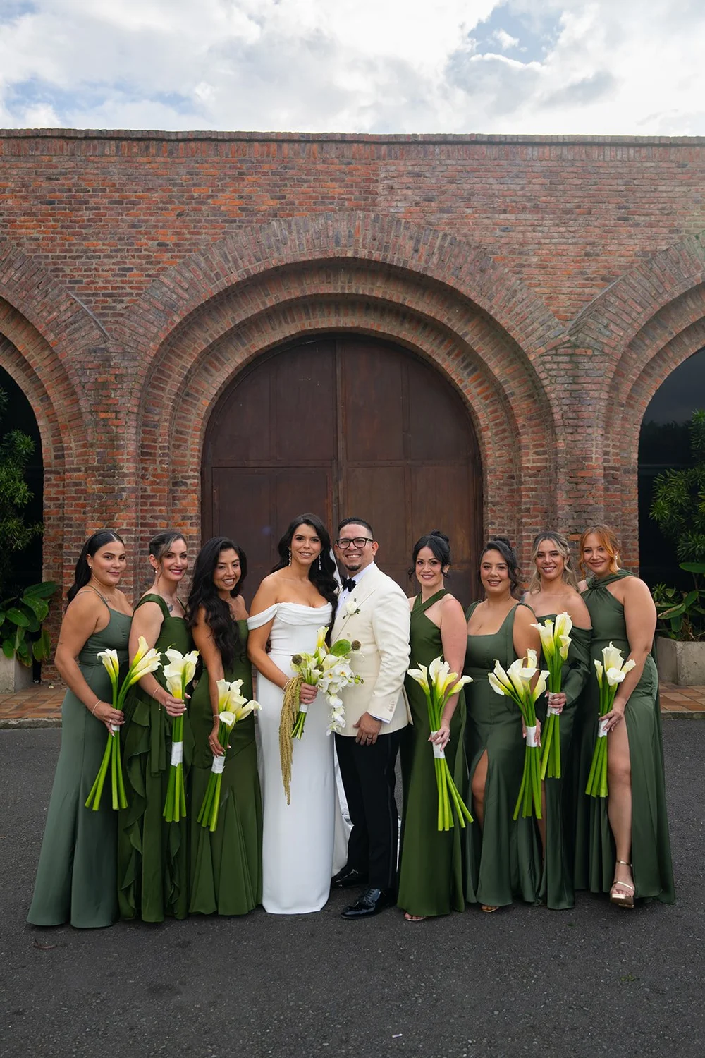 Krystine with her bridesmaids in green holding white flowers in front of a brick archway