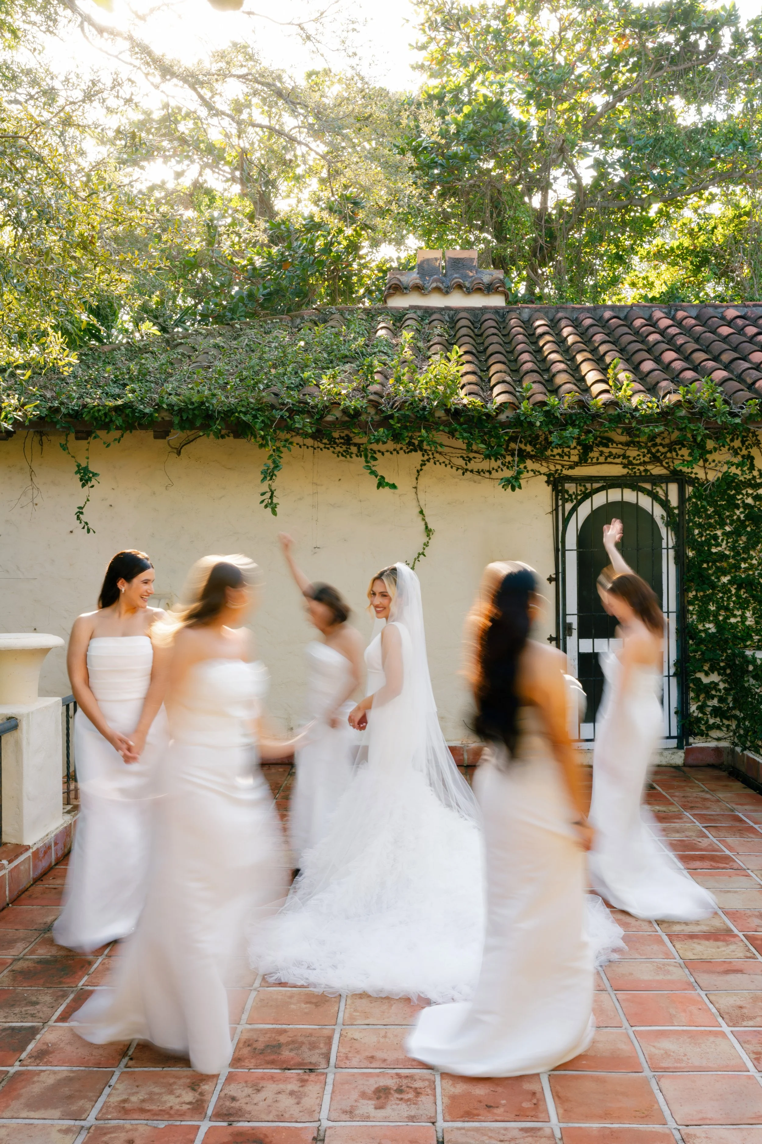 A bride surrounded by women in white dresses dancing outdoors, with trees and a tiled roof in the background.