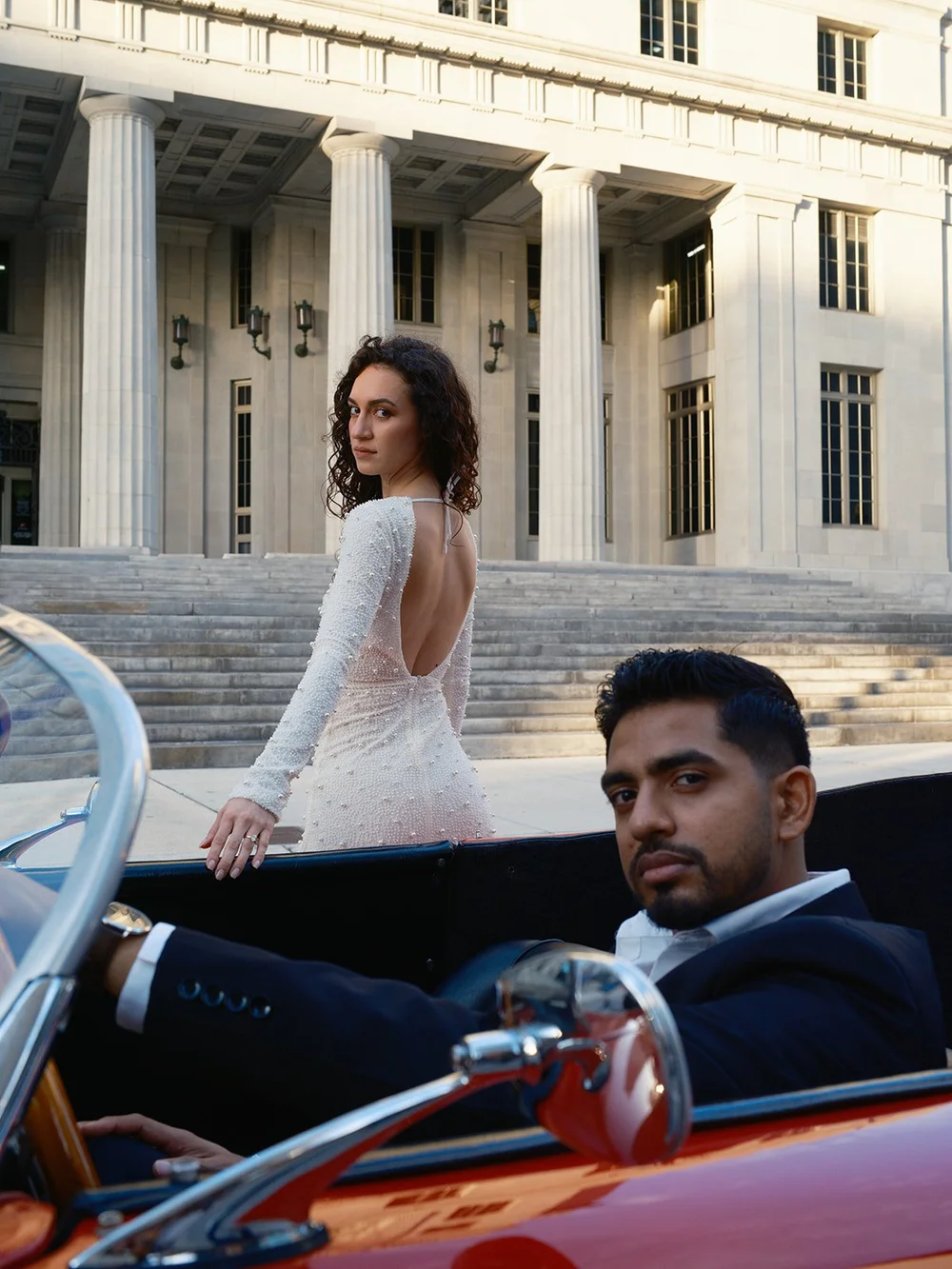 Bride standing in front of the courthouse steps while the groom sits in the driver's seat of the red convertible