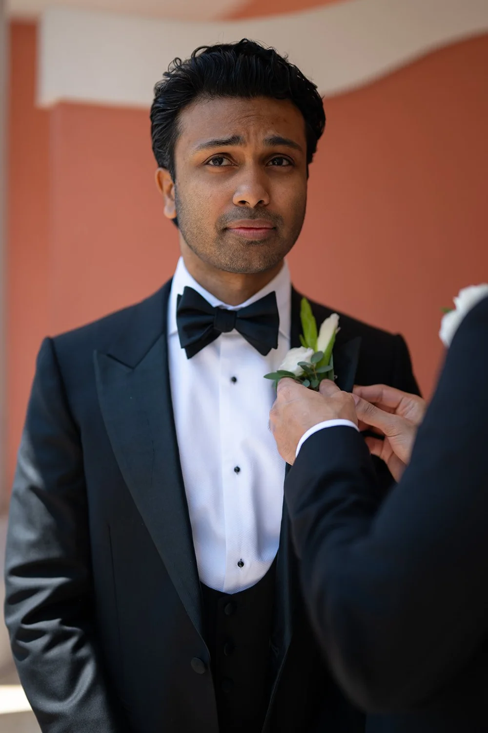 Jacob in his tuxedo with a white boutonniere being pinned to his lapel, with a pink wall behind him