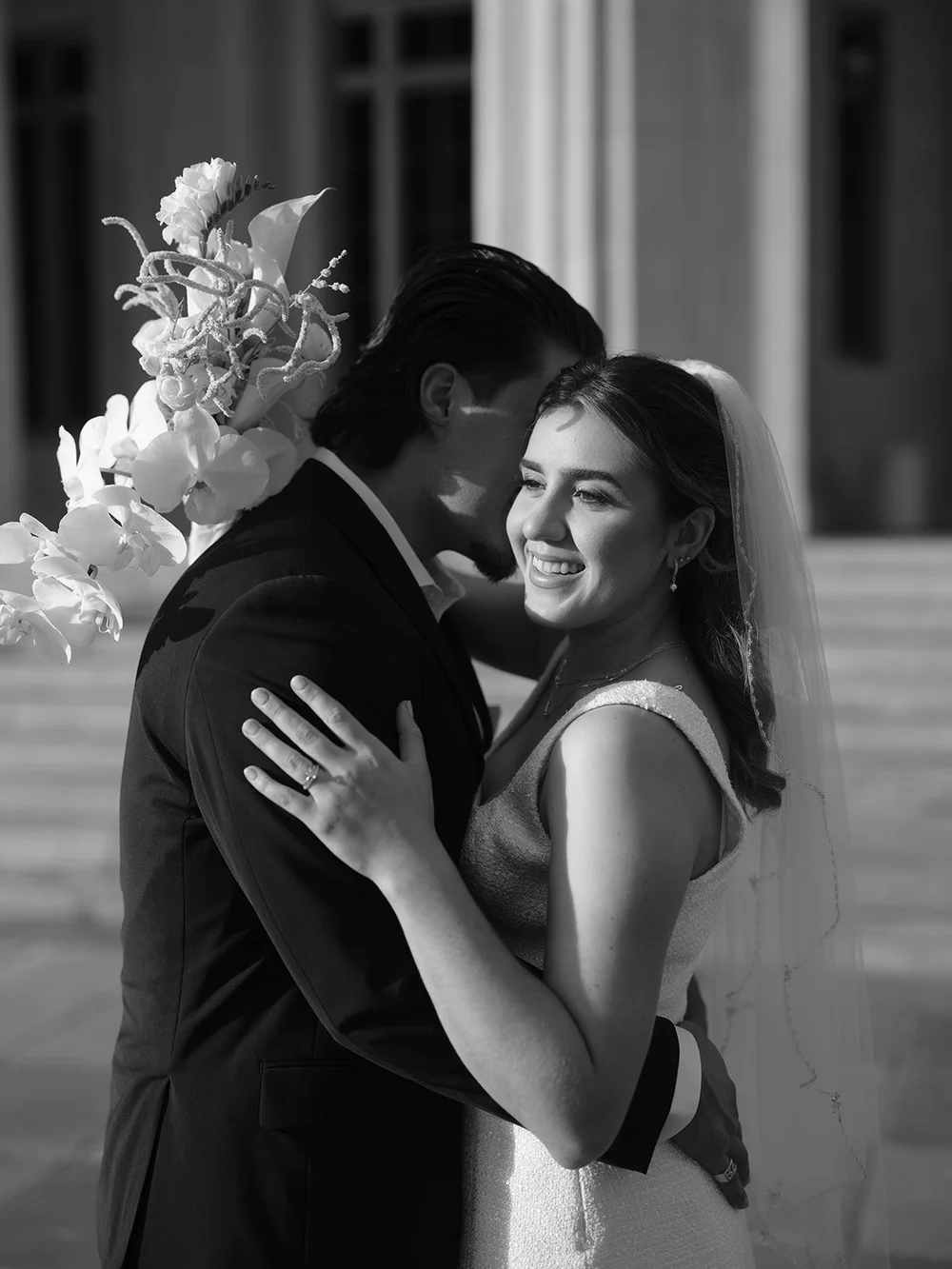 Black and white image of the groom kissing the bride's cheek while she smiles, white flowers above them