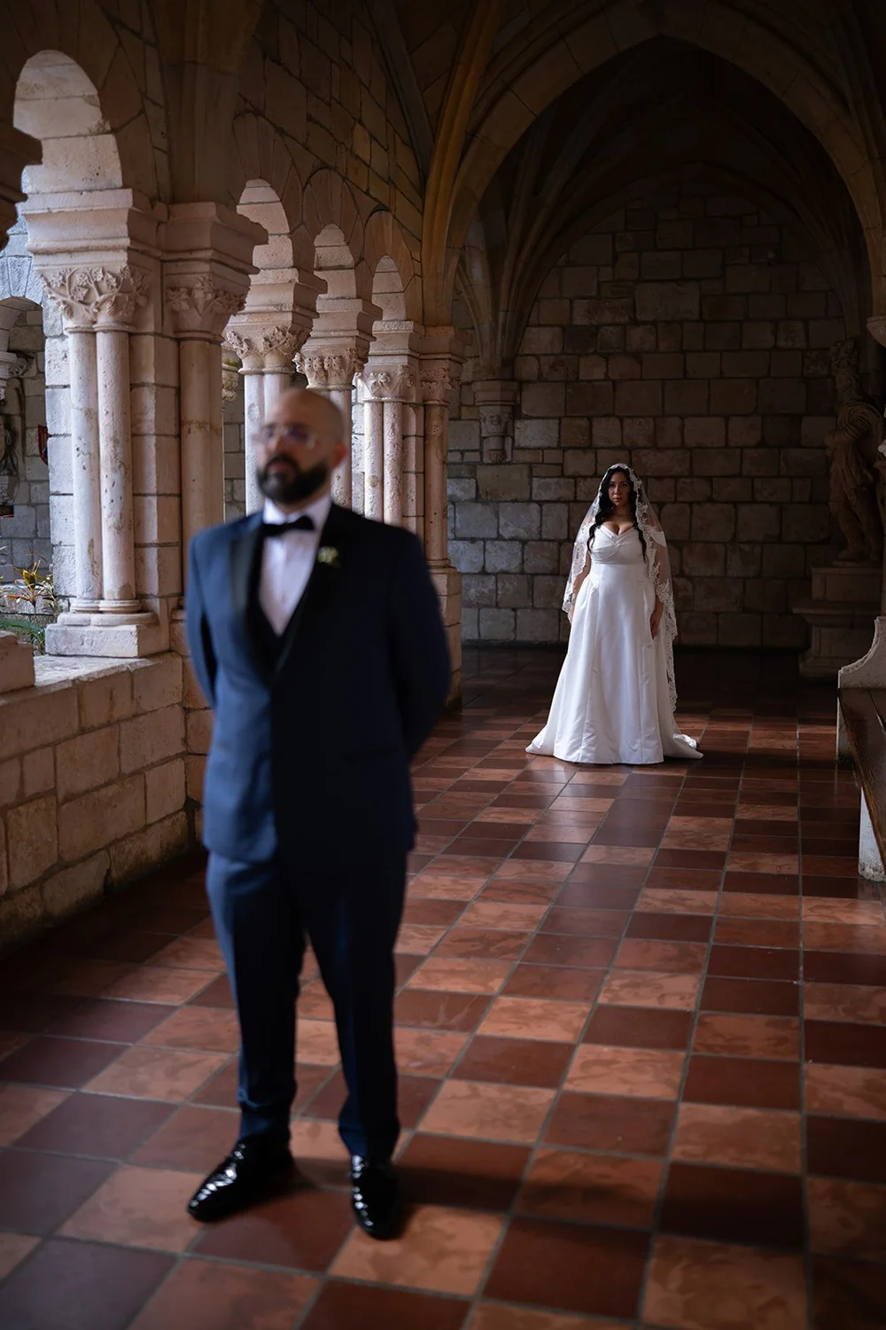 Christian in a navy tuxedo waiting in the cloister while Maria stands far behind him under the stone arches