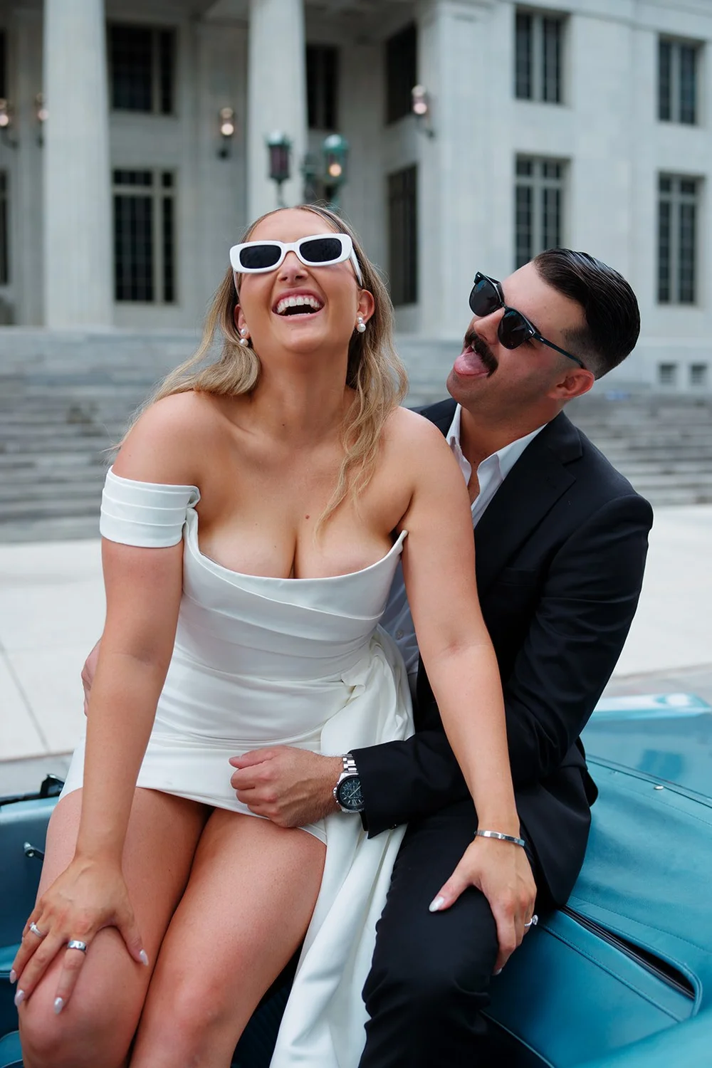 Rachel laughing in white sunglasses as Austin holds her on the hood of the Mustang