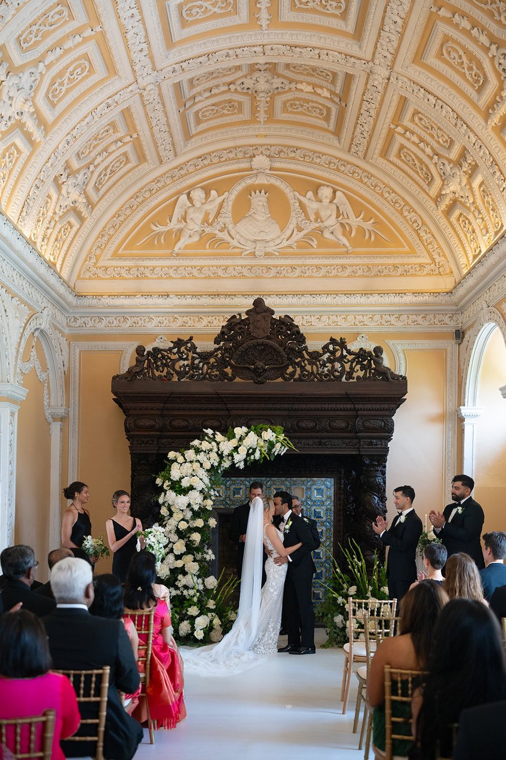 Ellen and Jacob at a carved dark altar framed by white flowers, with guests seated in the ornate hall