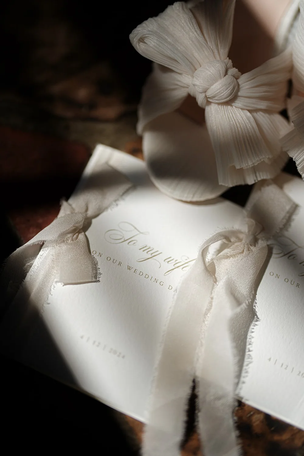 A wedding invitation tied with a ribbon next to white bow shoes in a patch of sunlight