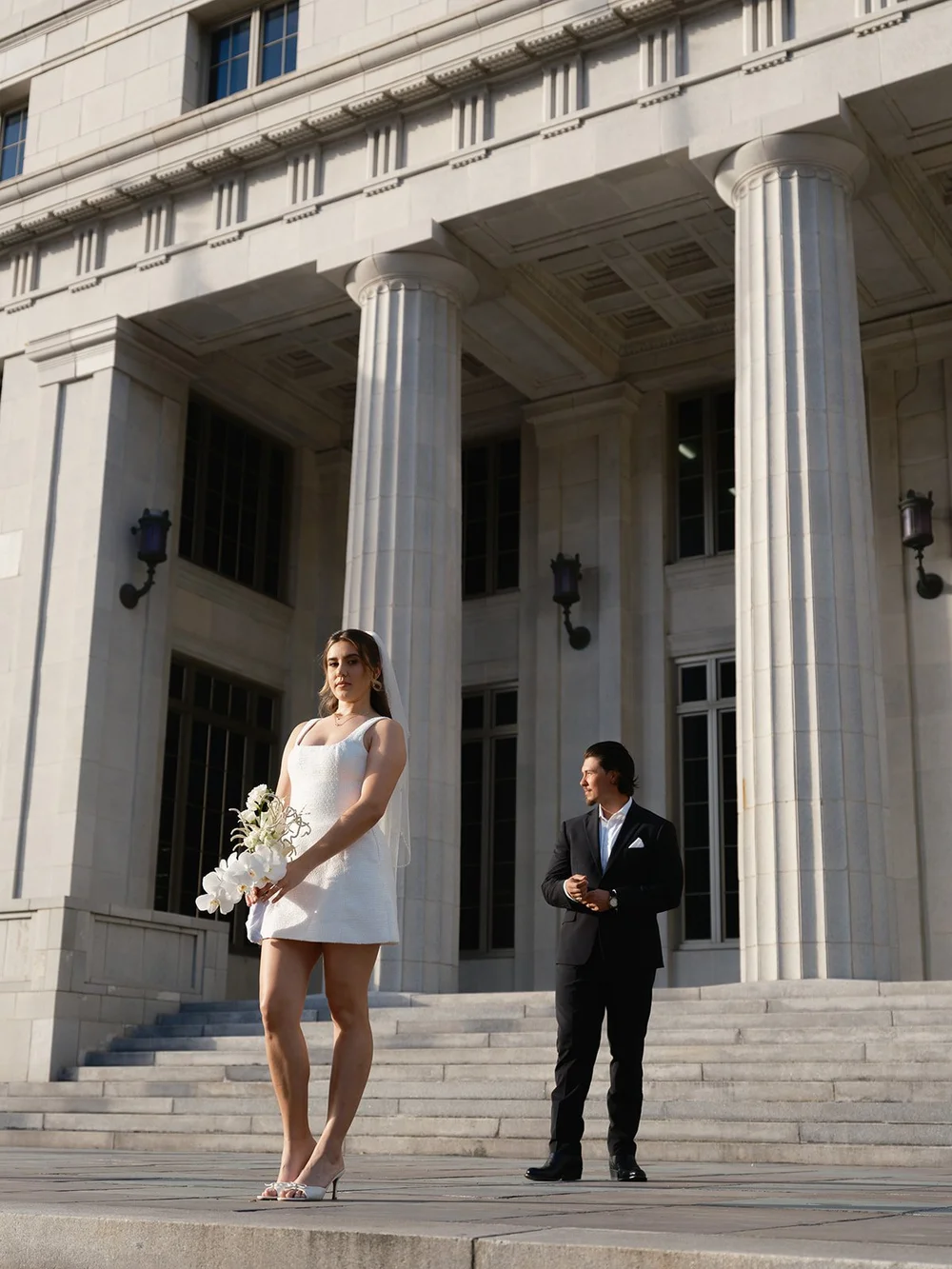 Bride in a short white dress and groom in a dark suit standing apart on the courthouse steps between two tall columns