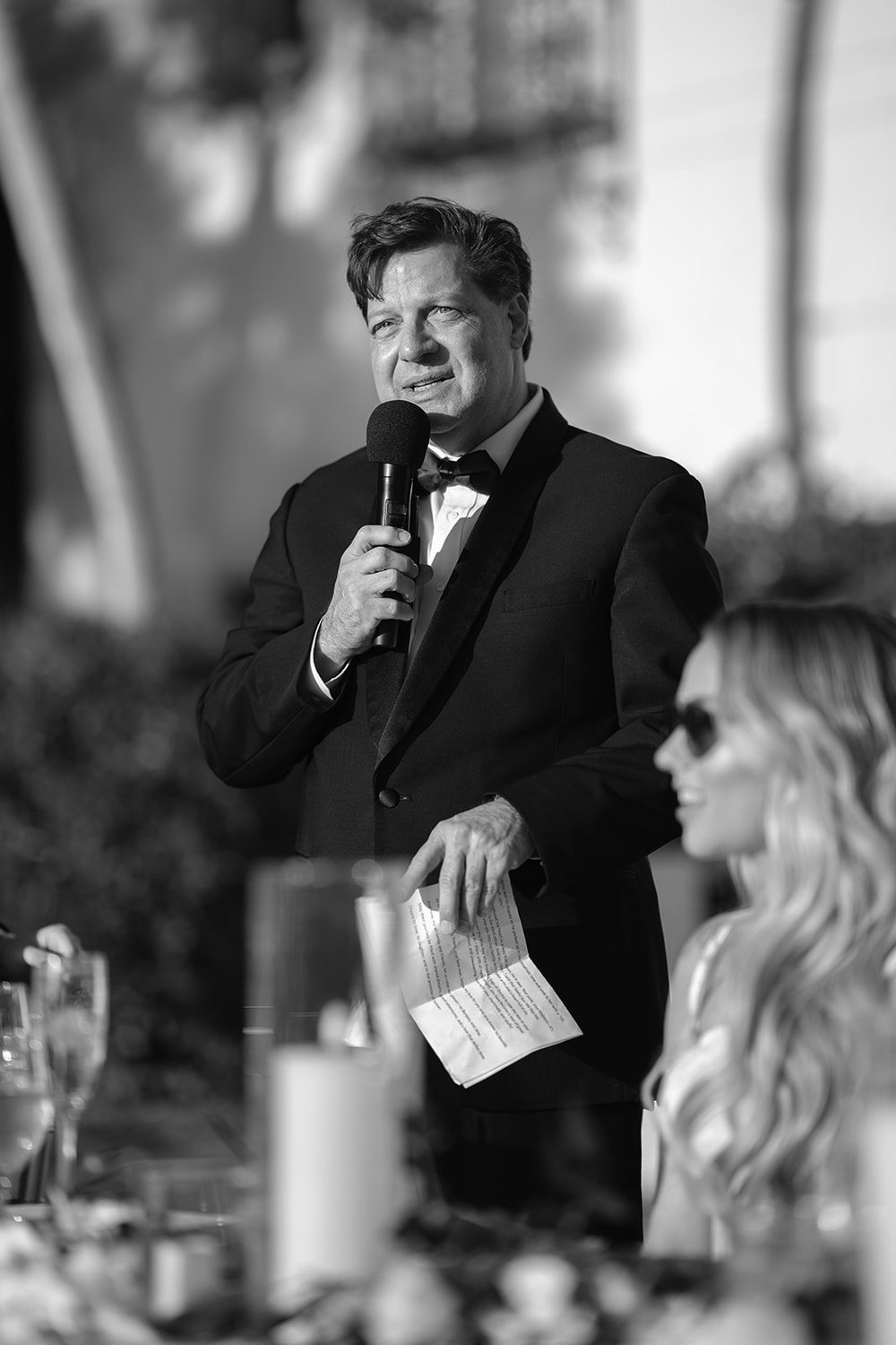 Black and white photo of a man giving a toast with a microphone while a woman in sunglasses listens