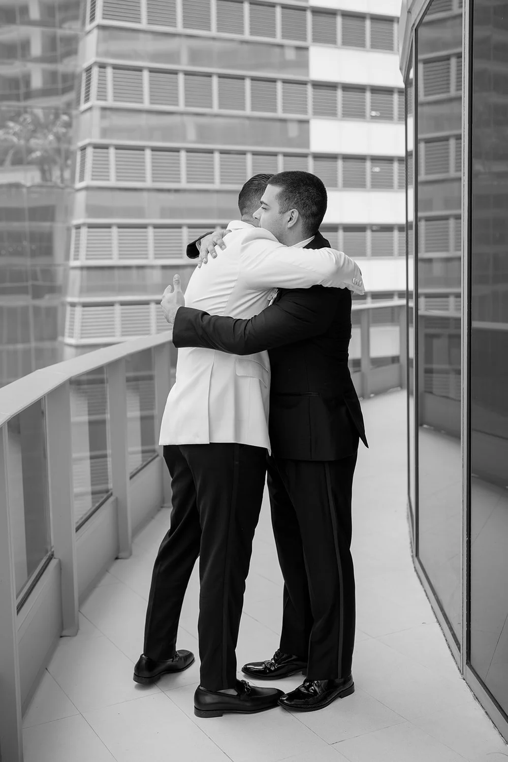 Two men in a hug on a balcony with downtown towers behind them, in black and white