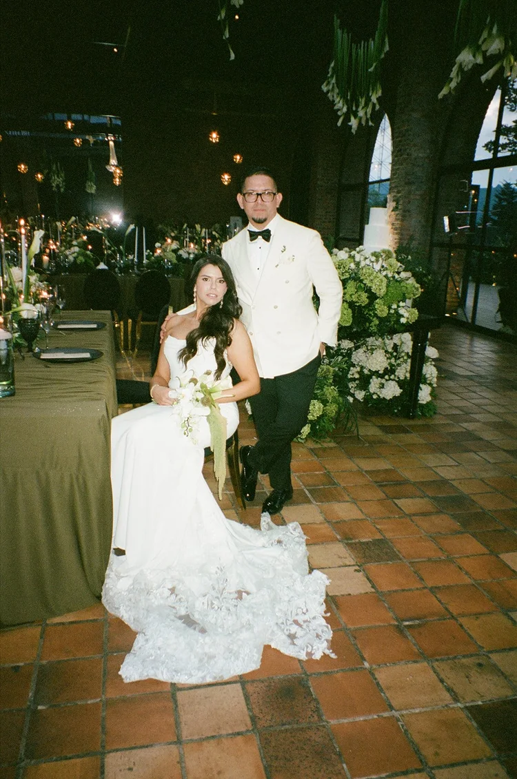 Film photo of Krystine seated in her gown with Douglas standing beside her in the brick reception room at Voilà Eventos