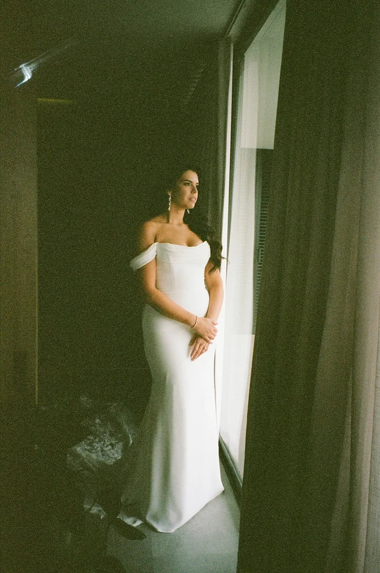 Film photo of Krystine standing in a window in her wedding gown with dark curtains beside her
