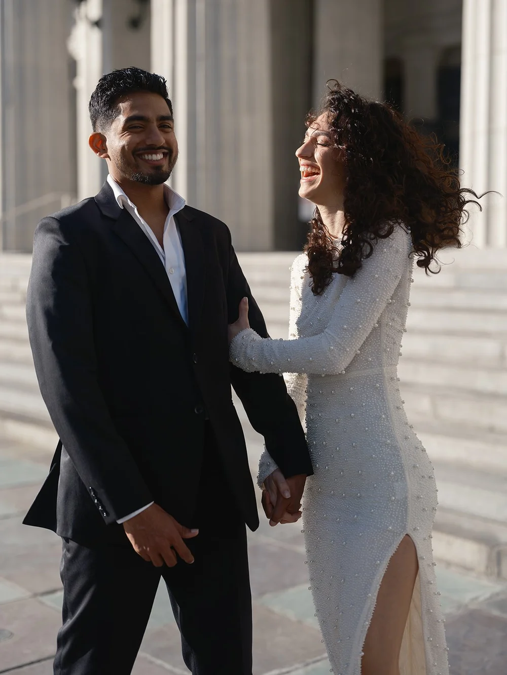 Black and white photo of the groom smiling and the bride laughing as they stand in front of the courthouse columns