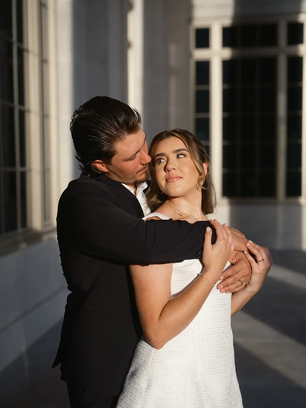 Groom hugging the bride from behind in warm light beside the courthouse's tall windows