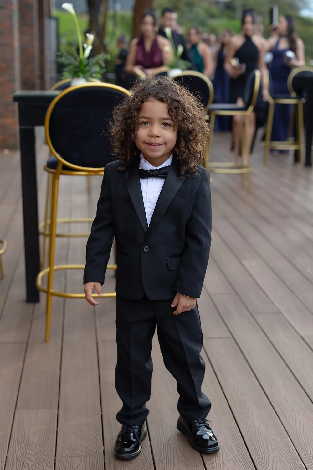 A young boy in a black tuxedo standing on the wooden deck