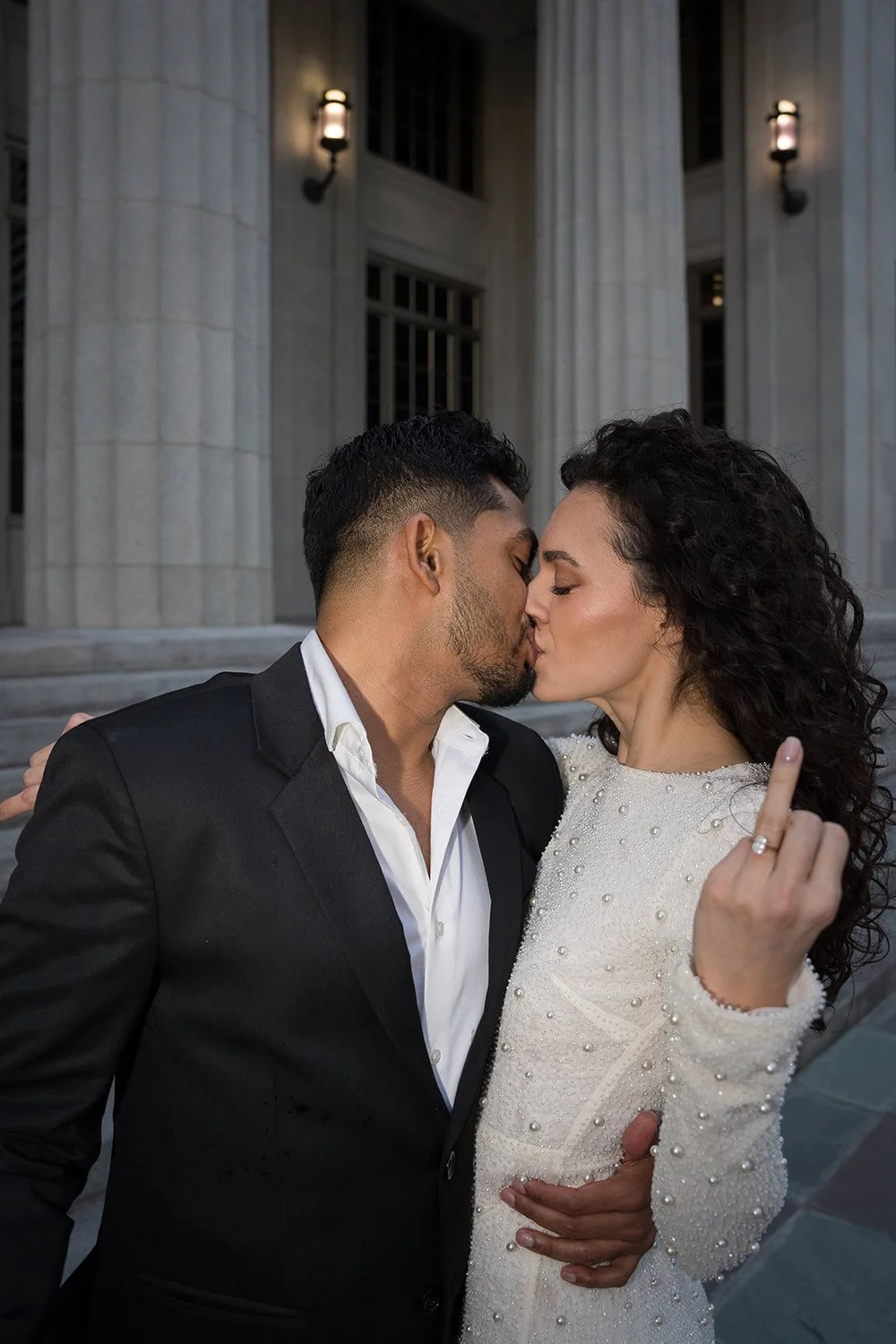 Black and white photo of the groom kissing the bride at dusk in front of courthouse columns and lamps