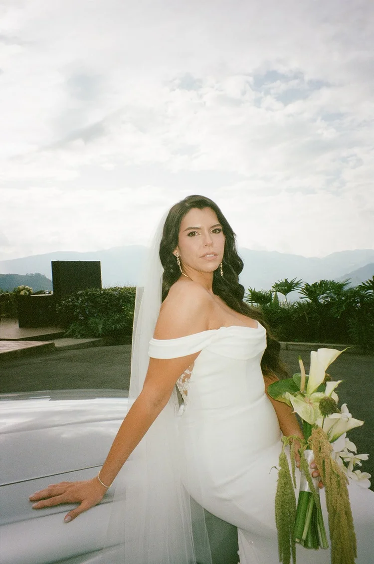 Film portrait of Krystine in an off-the-shoulder gown and veil leaning on a white car with a white calla lily bouquet