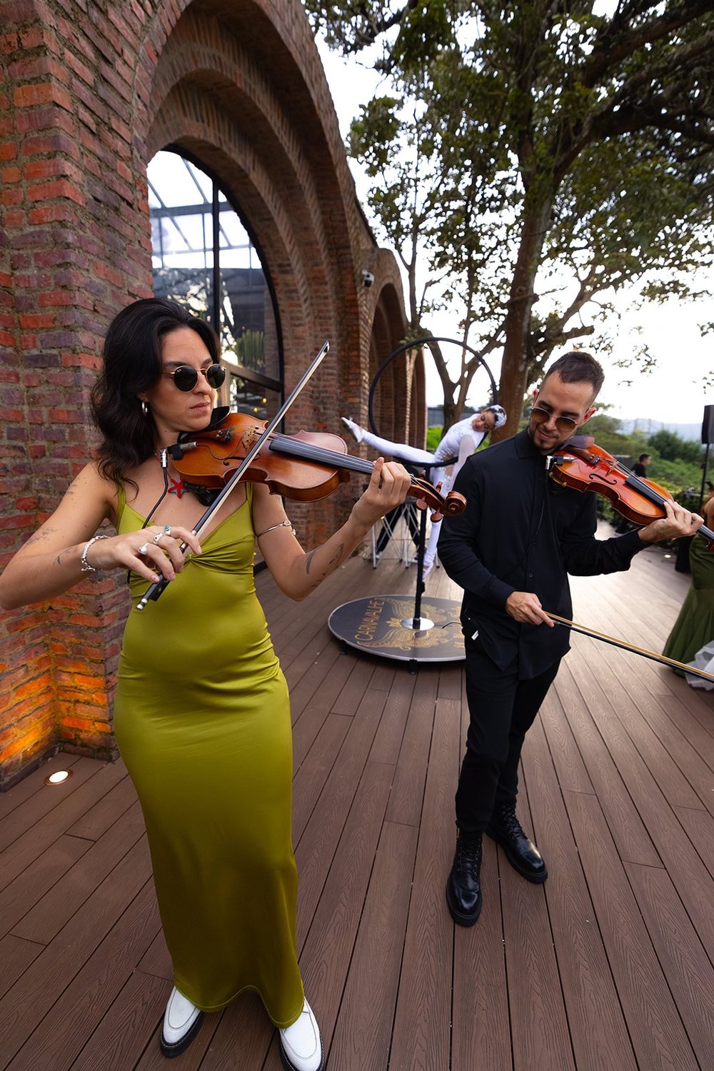 Two violinists playing on the wooden deck in front of a brick arch