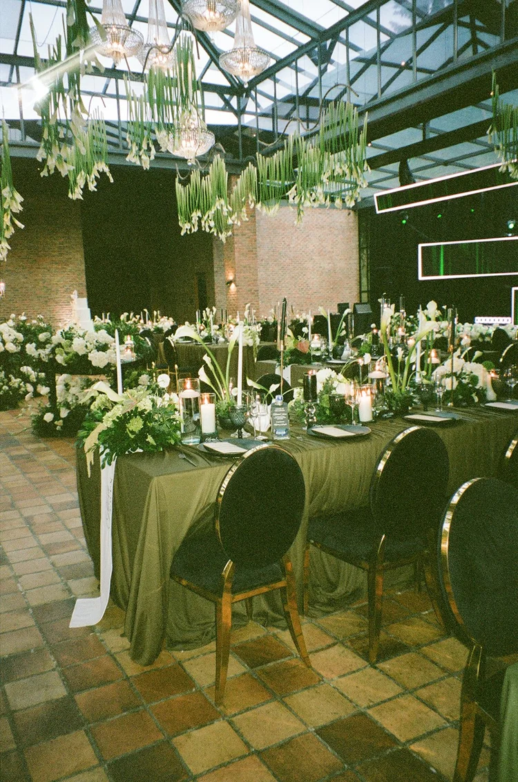 Film photo of a long reception table with green linens, candles and white flowers under a glass ceiling