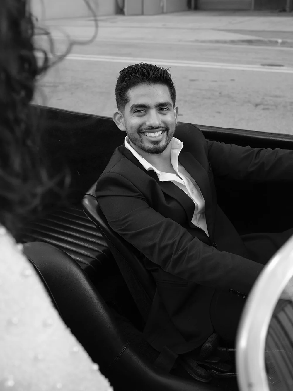 Black and white photo of the groom smiling at the camera from the seat of the red convertible