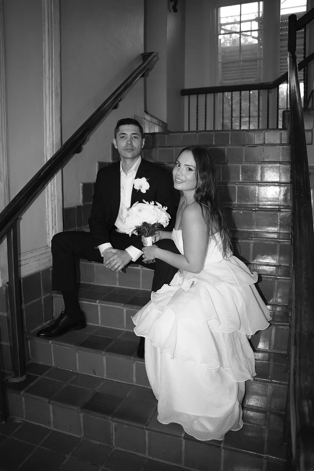 Black and white image of David and Chris sitting on a staircase, her tiered dress spread across the steps