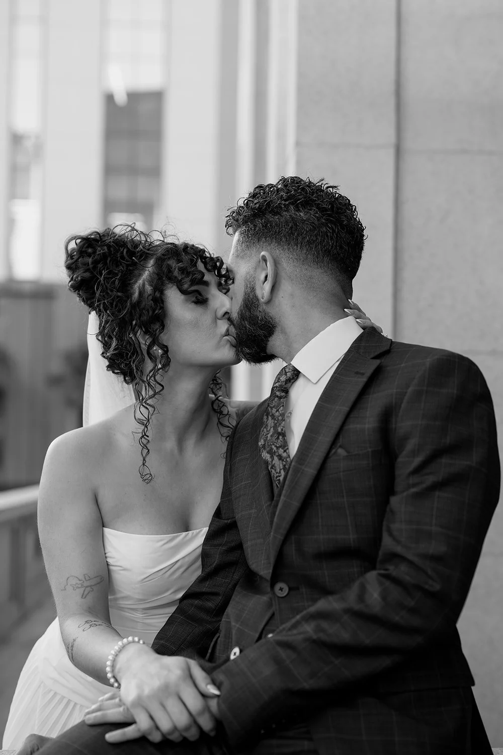 Black and white photo of the bride and groom kissing as they sit together beside a courthouse column