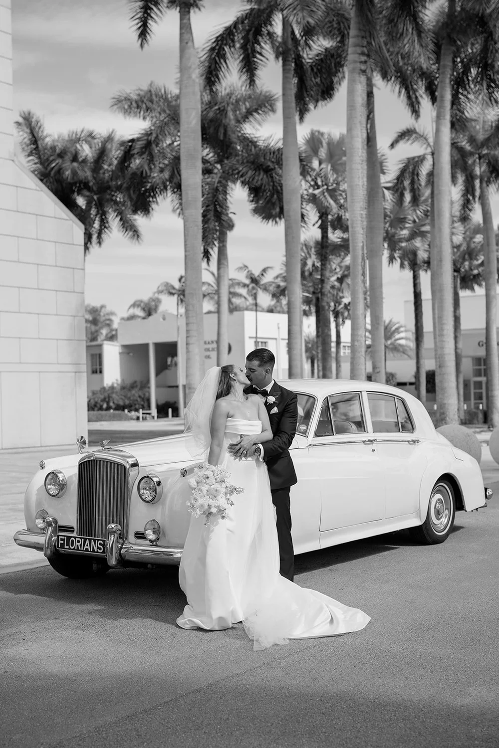 Rachel and Austin standing next to a vintage white car under palm trees, in black and white
