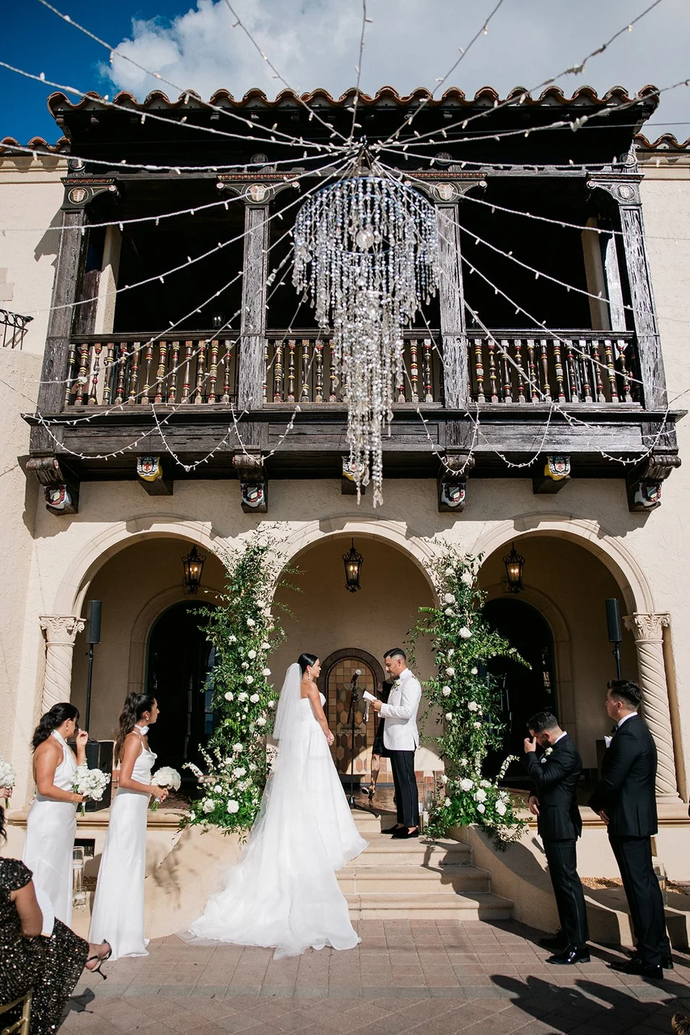 Steph and Ricky’s ceremony at the arched entrance of the Powel Crosley Estate house in Sarasota, with the wooden balcony, string lights and chandelier above them