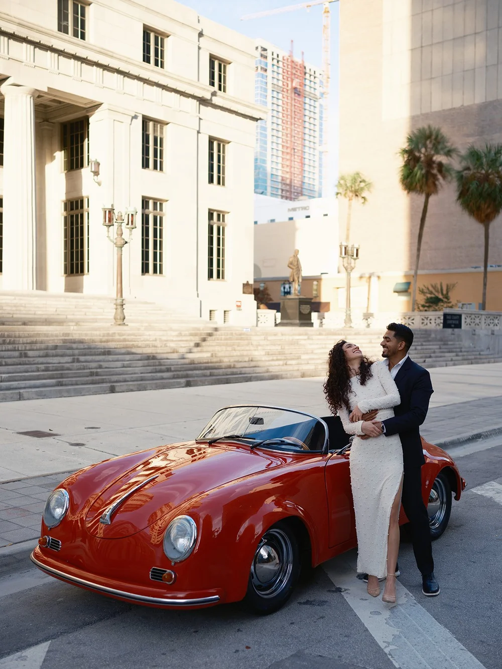 Bride and groom standing behind a red vintage convertible on a downtown street with courthouse buildings and palm trees behind