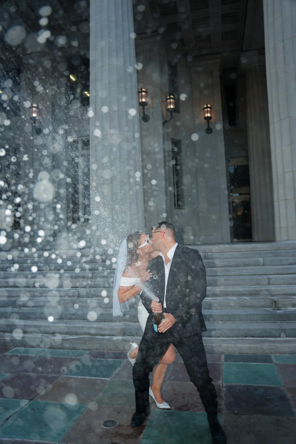 Rachel and Austin kissing in a spray of champagne in front of the courthouse columns