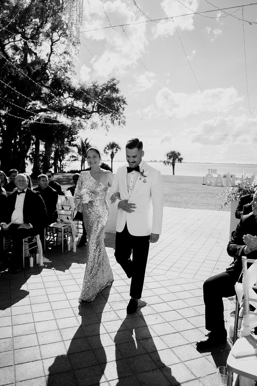 Black and white photo of Ricky in a white dinner jacket walking with a woman in a sparkly gown across the sunlit terrace past seated guests, with the water in the background
